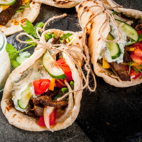 Three beef gyros with tzatziki sauce, cucumbers, tomatoes, and onions on a dark surface.