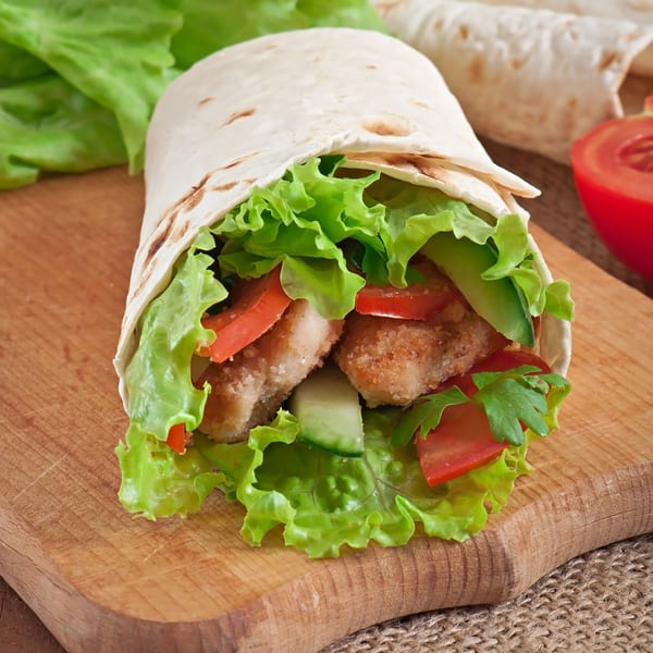 A chicken wrap with lettuce, tomatoes, and cucumbers, on a wooden cutting board.