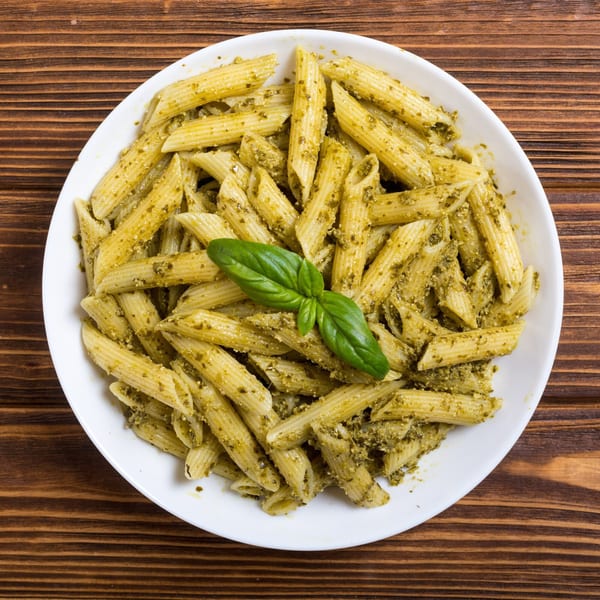 Penne pasta with green pesto sauce and a fresh basil leaf in a white bowl on a wooden table.