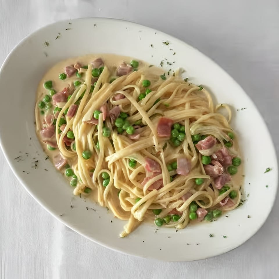Creamy pasta with peas and ham on a white oval plate, garnished with herbs.